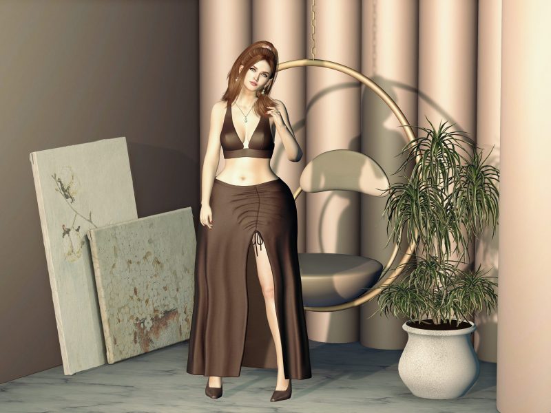 Second Life fashion avatar in a brown crop top and long skirt, standing beside a round hanging chair with a relaxed smile.
